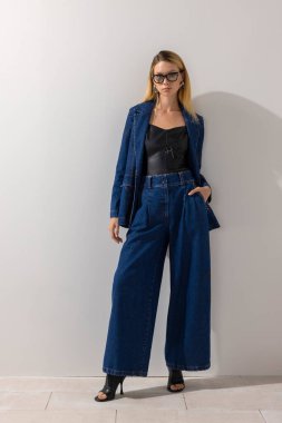 Büyük beden kot elbise ve deri bluz giyen modern stil sahibi kadın minimalist stüdyoda beyaz duvara yaslanıyor. Eğilim odaklı moda, temiz arkaplan ve kopyalama alanı.