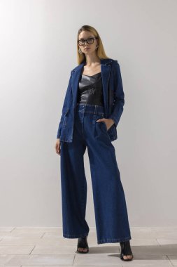 Modern kot elbise ve gözlük takan kendine güvenen şık bir kadın, beyaz arka planlı minimalist bir stüdyoda poz veriyor. Modern moda, zarafet ve kopyalama alanı.