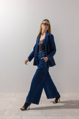 Büyük beden kot ve deri bluzlu modaya uygun bir kadın, minimalist stüdyoda beyaz arka planla güvenle yürüyor. Modern yaşam tarzı, moda eğilimleri ve kopyalama alanı.