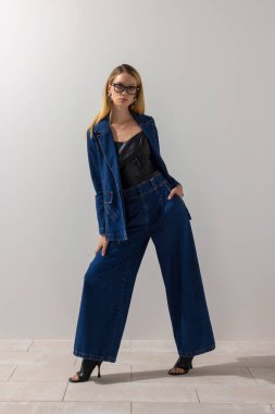 Aşırı büyük kot elbise ve deri bluz giyen şık modern bir kadın minimalist stüdyoda beyaz duvara karşı güven içinde poz veriyor. Eğilim odaklı moda, temiz arkaplan ve kopyalama alanı.