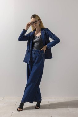 Kendine güvenen manken, minimalist stüdyoda büyük beden kot elbise ve deri bluz giyerken gözlük ayarlıyor. Modern iş gündelik tarzı, yaşam tarzı ve kopyalama alanı.