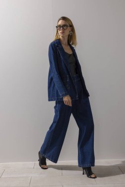 Kendine güvenen, kot ceket ve geniş bacak kotuyla yürüyen, minimalist stüdyoda gözlük takan modern bir kadın. İş modası, yaşam tarzı, liderlik ve kopyalama alanı.