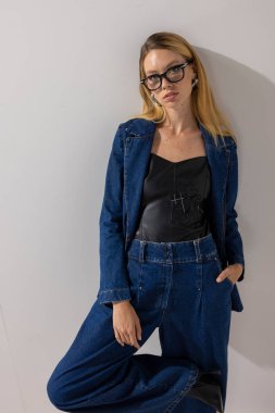 Kot ceket giyen şık sarışın kadın, geniş paçalı kot pantolon ve gözlük, minimalist stüdyoda beyaz duvara karşı poz veriyor. Moda, yaşam tarzı, fotokopi alanı ile iş gündelik konsepti.