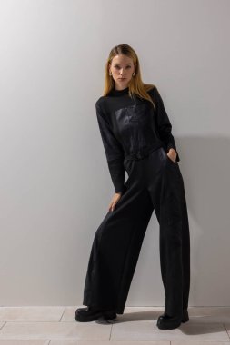 Modern siyah tulum giyen modacı kadın minimalist stüdyoda beyaz duvara yaslanıyor. Güç sargısı, çağdaş yaşam tarzı, editoryal moda, ve kopyalama alanı.
