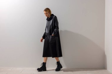 Modern siyah bir elbise ve botla yürüyen bir kadının çağdaş moda görüntüsü. Profilde minimalist bir stüdyo duvarına karşı çekilmiş. Temiz çizgiler, hareket ve editoryal biçim.