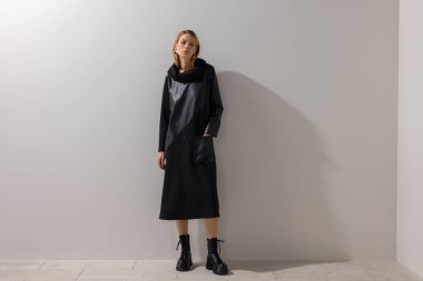 Tarafsız bir stüdyo duvarına karşı duran, modern siyah bir elbise ve çizme giyen genç bir kadının tam boy moda portresi. Temiz kompozisyon, minimalist tarz ve editoryal moda..