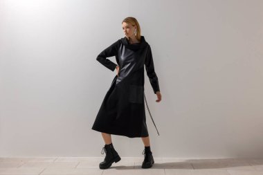 Modern siyah bir elbise ve botlarla, temiz bir stüdyo duvarına karşı duran bir kadının editörlük modası. Kalın duruş, minimalist kompozisyon ve çağdaş biçim.