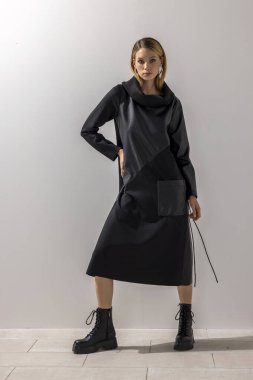 Siyah bir Midi elbisesi ve asker botu giyen bir kadının modern moda portresi, temiz bir stüdyo geçmişine karşı güven içinde poz veriyor. Güçlü minimalist tasarım, çağdaş siluet ve editoryal moda..