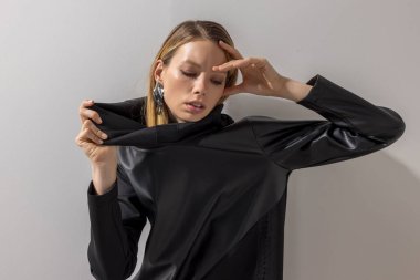 Yayın tarzı moda portresi, siyah deri kapüşonlu, anlamlı poz ve küpe takan, doğal ışıkta, nötr bir duvara yaslanmış genç bir kadın..