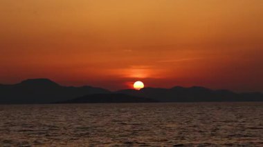 Dağ siluetleri ve turuncu gökyüzü ile sakin denizin üzerinde gün batımı. Ufkun altında batan güneş dramatik, barışçıl ve sinematik bir arka plan yaratır..