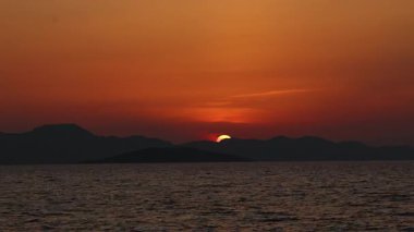 Güneş ufkun altında kaybolurken sakin deniz ve dağ siluetleri üzerinde gerçek zamanlı gün batımı. Sıcak turuncu gökyüzü alacakaranlıkta soluyor, sakin bir sinematik son yaratıyor, duygusal hikaye anlatımı ve doğa arka planı görselleri için ideal..