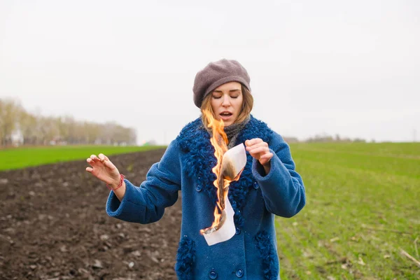 Desperate angry girl in blue coat hold burning paper with fire at open cold windy field, female frustration and sadness, start life from new page
