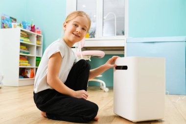 İlkokul çağındaki sarışın kız hava nemlendirici beyaz kutu, sağlık hizmetleri ve modern cihazlarla evinde rahat ediyor.