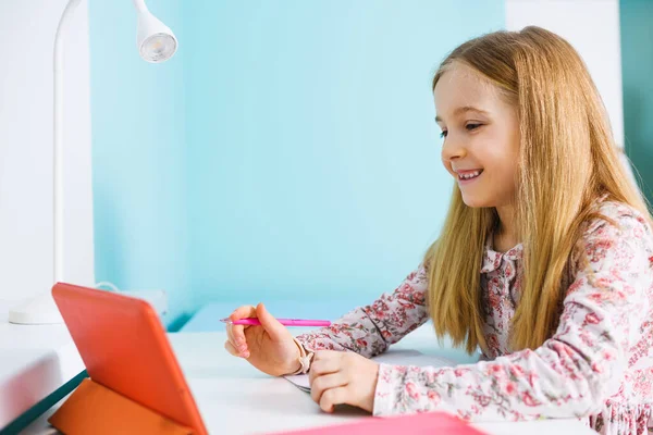 小学生の女の子は机に座って ペンと紙のノートブックでタブレット画面のために笑顔 家の隔離中のリモート研究 — ストック写真