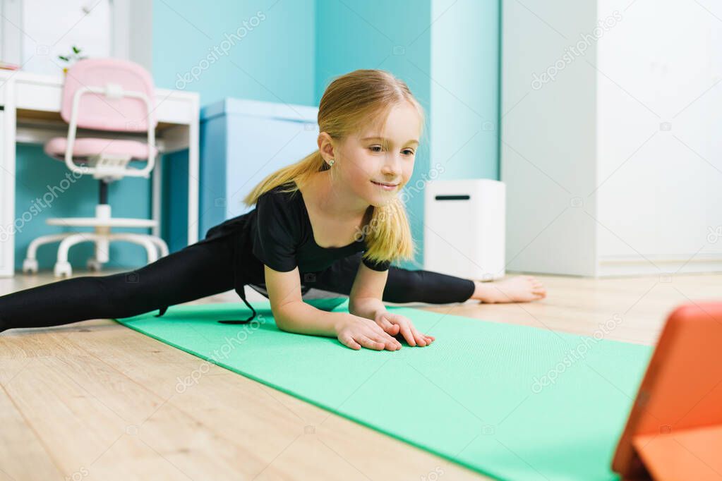 Primary school age sportive gymnast girl in black leotard stay in twine pose and look at tablet screen during online training at home