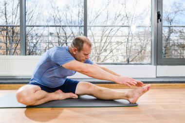 Orta yaşlı zinde bir adam ışık stüdyosunda yere oturur ve ellerini ve ayaklarını uzatır, yoga egzersizleri yapar.