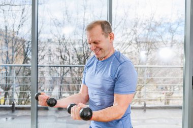 Orta yaşlı, formalı, mavi tişörtlü, spor salonundaki dambıllı, panoramik pencereli bir adam.