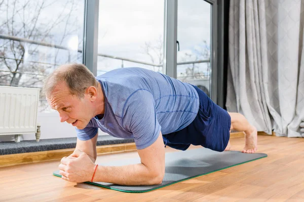 Yaşlı formda sporcu, kalas yoga pozu veriyor. Odadaki paspasın üzerinde duruyor. Yüzü yakın, sağlıklı bir yaşam tarzı.
