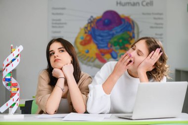 İki genç kız öğrenci okul konferansında kendilerini sıkıcı ve yorgun hissediyorlar. Modern sınıf masasında dizüstü bilgisayar ve DNA modeliyle esniyorlar.