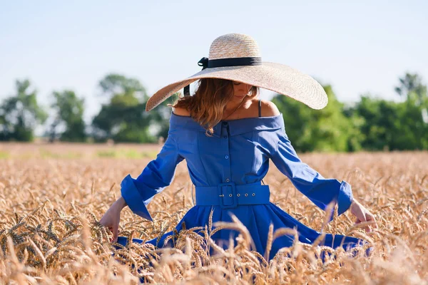 Romantik moda kızı buğday tarlasında duruyor, kemerli ve geniş kenarlı hasır şapkalı mavi bir elbise giyiyor.