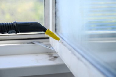 Bir buharlı temizleme hortumunun pencerenin çerçevesini odanın içinden temizlerken yakın plan görüntüsü. Camda görünür buhar ve yansıma var. Parlak gün ışığı ve bulanık yeşillik.