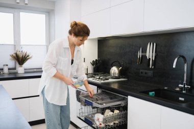 Genç bir kadın, açık bir bulaşık makinesine parlak, modern bir mutfakta bir kase koyuyor. Günlük ev rutini, soba ve tezgahın yanında durmak, çağdaş iç mekan, doğal iç mekan ışığı.