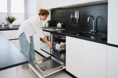 Genç bir kadın, modern bir minimalist mutfakta bardakları ve bulaşıkları açık bir bulaşık makinesine koyuyor. Sıradan ev işleri, ayakta duruş, çağdaş iç mekan, doğal gün ışığı..