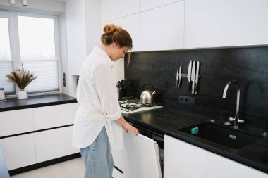 Genç bir kadın modern, parlak bir mutfakta bulaşık makinesini çalıştırmak için kontrol paneline basıyor. Günlük ev işleri, tezgah ve sobanın yanında durmak, çağdaş iç mekan, doğal iç mekan..