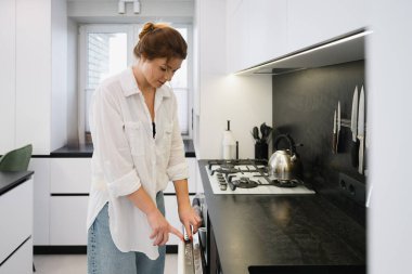 Genç bir kadın modern, parlak bir mutfakta bulaşık makinesini çalıştırmak için kontrol paneline basıyor. Günlük ev işleri, tezgah ve sobanın yanında durmak, çağdaş iç mekan, doğal iç mekan..