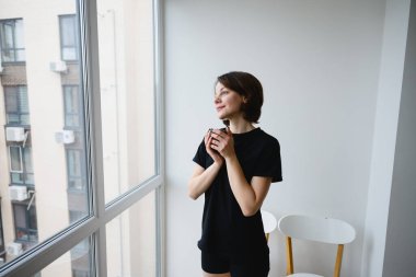 Şehir manzarasına pencereden bakarken elinde bir fincan kahve tutan düşünceli bir kadın. Sakin bir sabah anı, farkındalık, yaşam tarzı ve evde sakin bir yansıma..