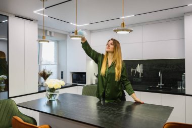 Modern bir mutfak içerisindeki bir lambanın tozunu silen bir kadın. Ev temizliği, günlük ev işleri, minimalist daire, doğal ışık..