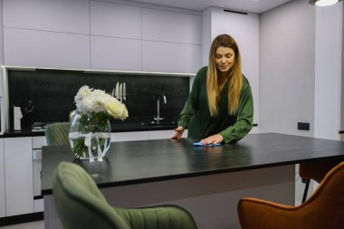 Yetişkin bir kadın modern bir ev içinde mutfak adasının tozunu bez parçasıyla temizliyor. Günlük temizlik rutini, minimalist daire, ev temizliği.