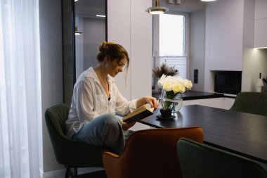 Masada oturan genç bir kadın modern bir dairede bir fincan kahveyle kitap okuyor. Sakin bir sabah rutini, rahatlama ve rahat bir ev hayatı..