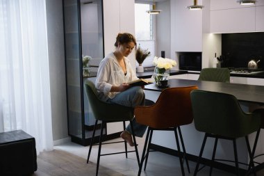 Genç bir kadın, modern bir dairede bir sandalyede oturmuş mutfak masasında kitap okuyor. Sakin boş zaman, rahat ev atmosferi ve sakin günlük yaşam tarzı..