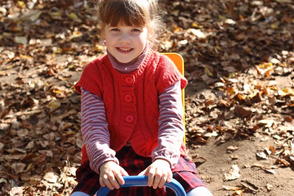 Petite jolie fille souriante s'assoit sur une scie à sous en bois sur une aire de jeux a — Photo