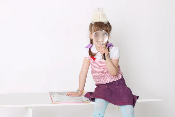 Little girl sits on bench with book and looks through magnifying — Stock Photo, Image