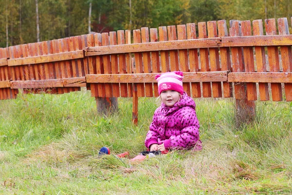 Маленькая красивая девочка сидит в траве возле оранжевого деревянного забора в ауте — стоковое фото