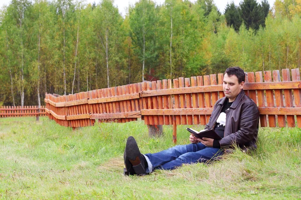 Mann liest an Herbsttag Buch auf Gras nahe orangefarbenem Holzzaun — Stockfoto