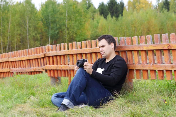 Man met de camera zit op gras in de buurt van Oranje houten hek aan herfst — Stockfoto