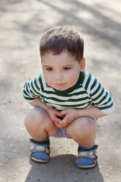 Happy little cute boy squats on asphalt in park at summer day. S — Stock Photo, Image