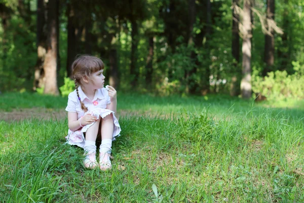 Pretty happy girl sits on green grass in forest and eats at sunn — Stock Photo, Image