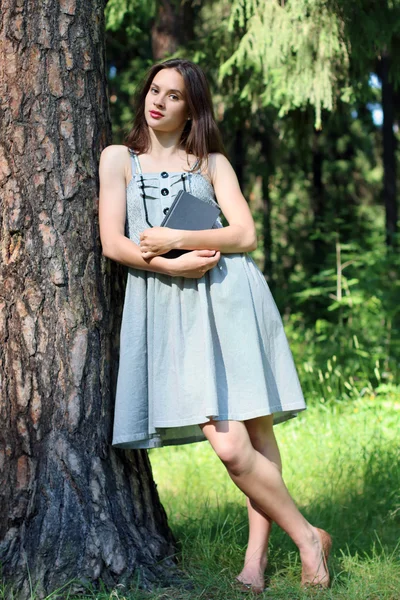 Hermosa chica en vestido de pie apoyado contra el árbol con libro — Foto de Stock