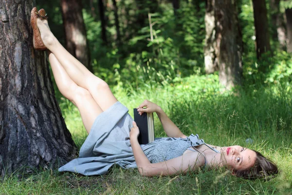 Beautiful girl in dress lying on grass under tree with book and — Stock Photo, Image
