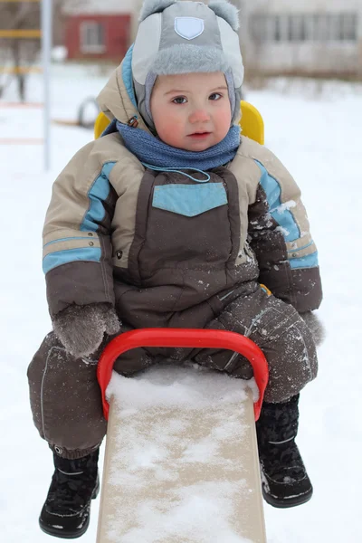 Küçük çocuk sıcak tulum giyen tahterevalli üzerinde kış gününde oturur. — Stok fotoğraf