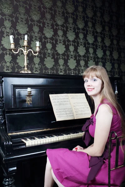 Mujer joven y bonita se sienta en la habitación cerca del piano, sonríe — Foto de Stock