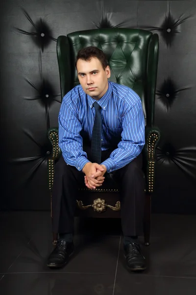 Serious young man in blue sits in leather armchair in room — Stock Photo, Image