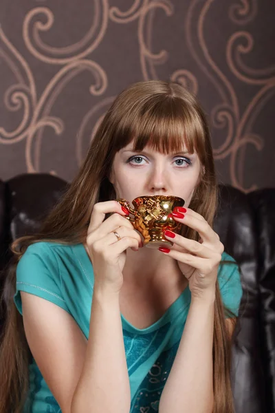 Young beautiful woman drinks coffee from gilt cup on couch — Stock Photo, Image