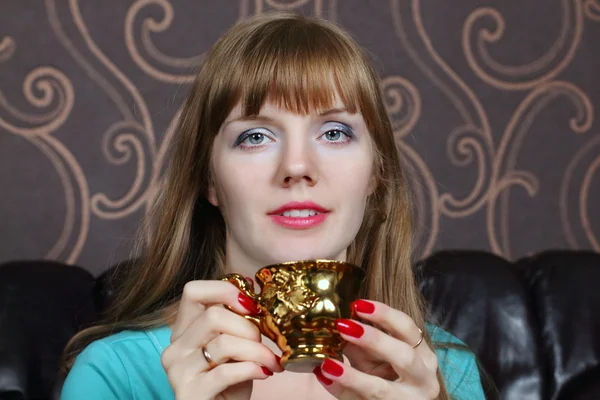 Young beautiful woman smiles and holds gilt cup on couch in room — Stock Photo, Image