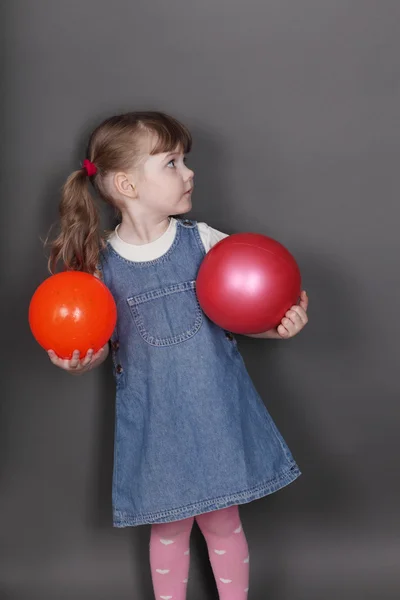 Pretty little girl in jeans dress holds two balls in grey studio — Stock Photo, Image