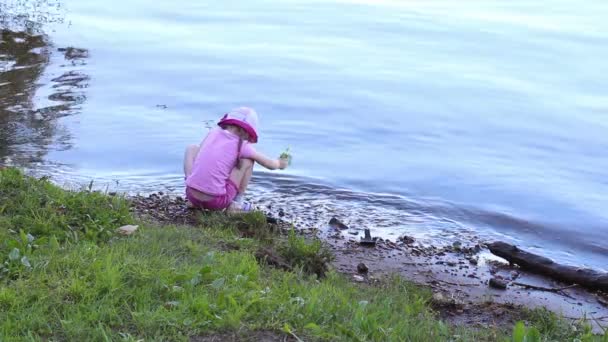 Обратная сторона маленькой симпатичной девочки в розовом, сидящей на берегу реки и смотрящей на воду — стоковое видео